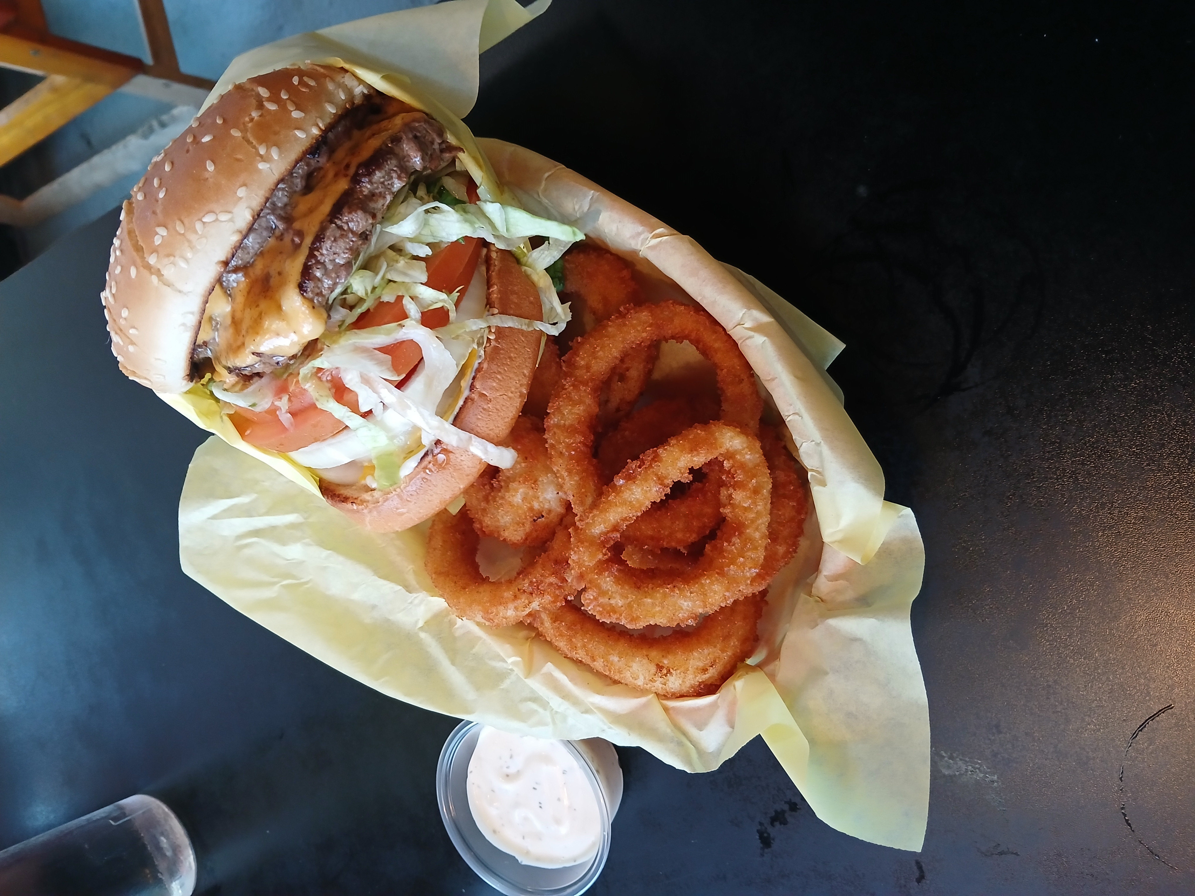 A double cheeseburger wrapped in paper and sitting behind a serving of onion rings inside of a plastic serving basket. Ranch sits to the left of the meal.