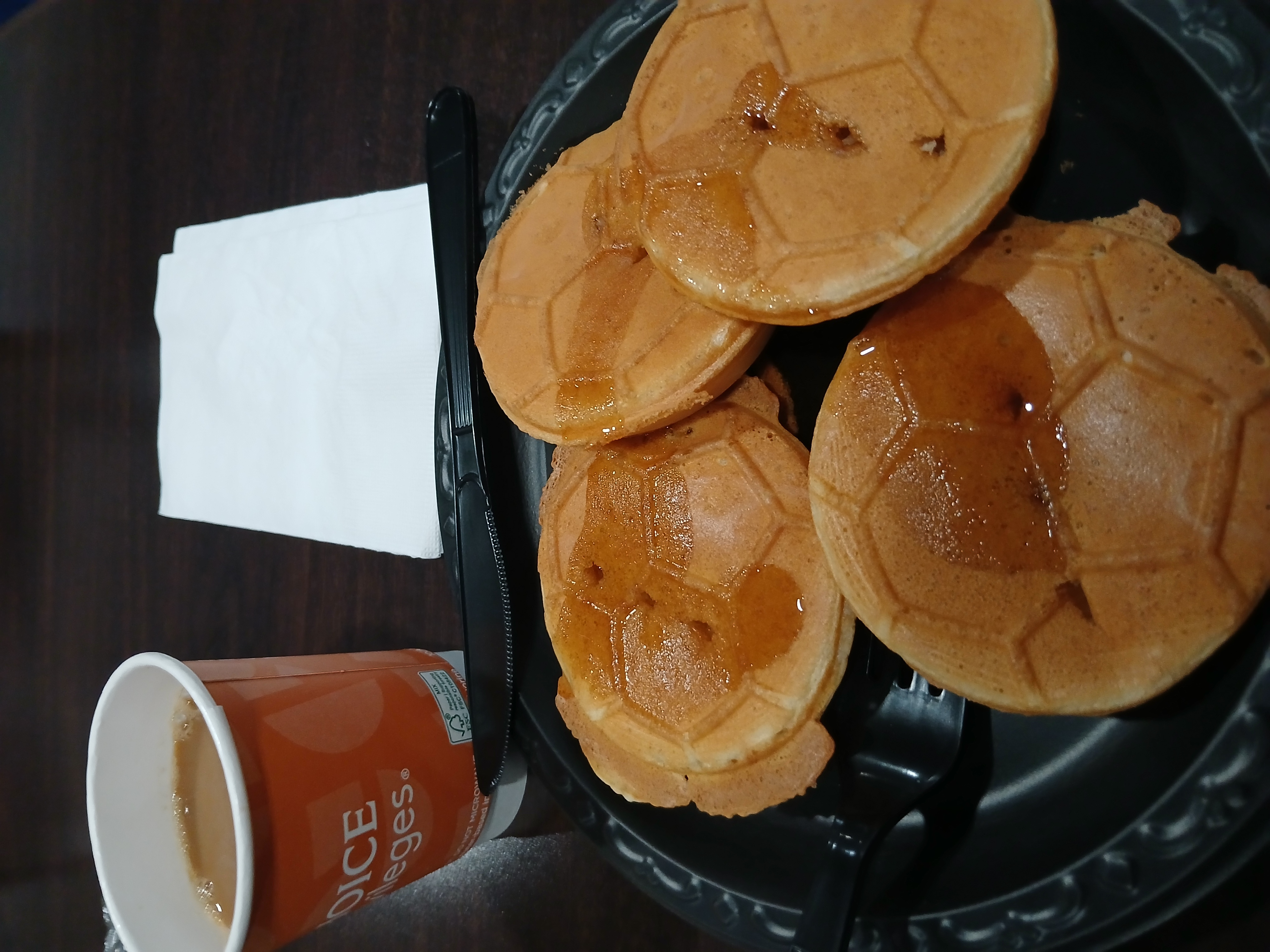 Crisp waffles with some trim left on the edges and slathered in syrup. The iron has cooked them into the impression of a soccer ball on the side facing the camera. A plastic fork and knife sit on the edge of the plate. A cup of coffee and stack of napkins sit on the side.