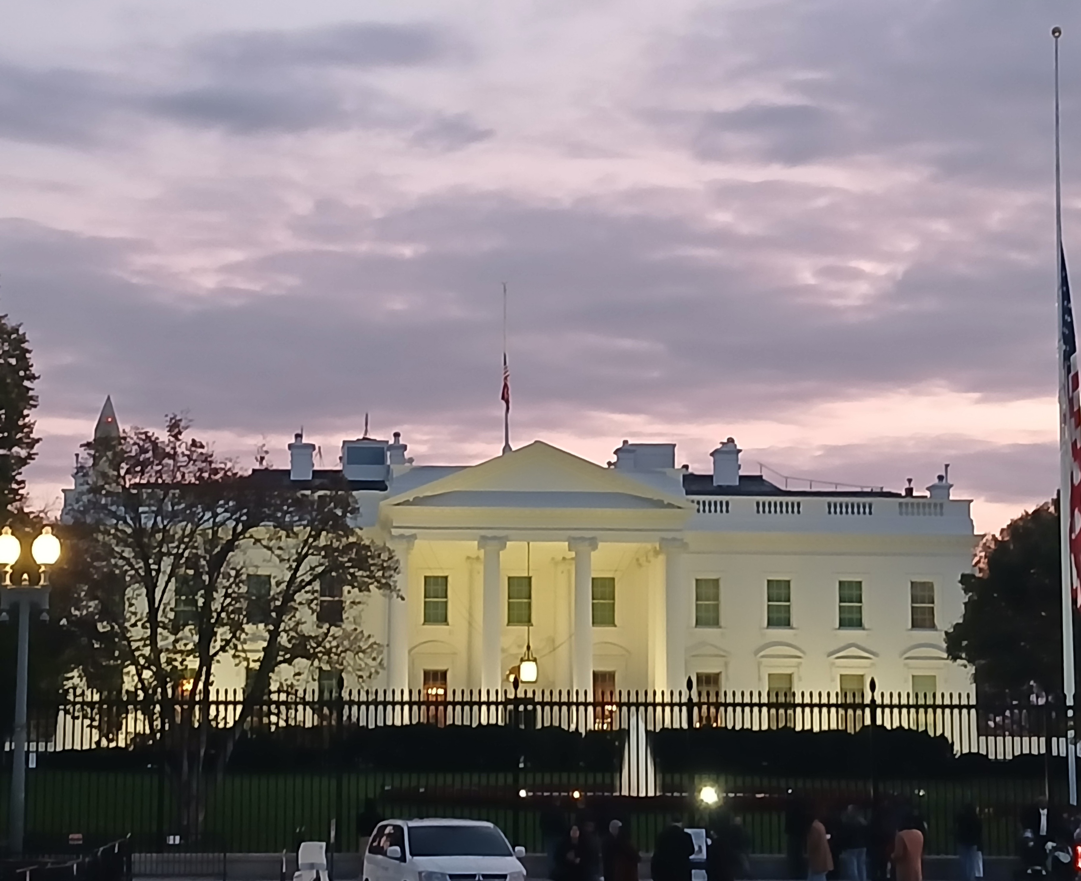The White House pictured in the evening. The US flag hangs drapped over a tall flag pole in the foreground.